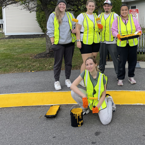 A group of students wearing yellow safety vests pose in front of the Westbrook Housing building displaying the painted sidewalk curbs they completed to enhance safety