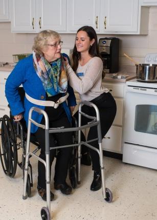 A student assists an elderly person move from their wheelchair to their walker